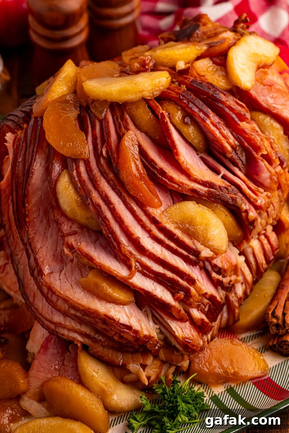 view of a large sliced spiral ham on platter with some apples on top and around with a red checkered towel behind it 