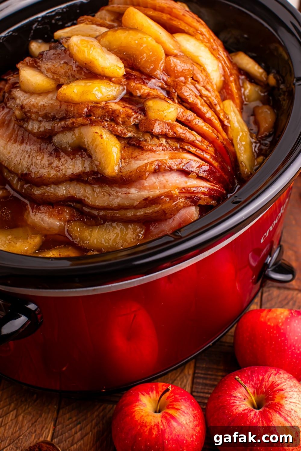 red slow cooker on a wooden table with a spiral ham with apples on top, and whole apples in front of table