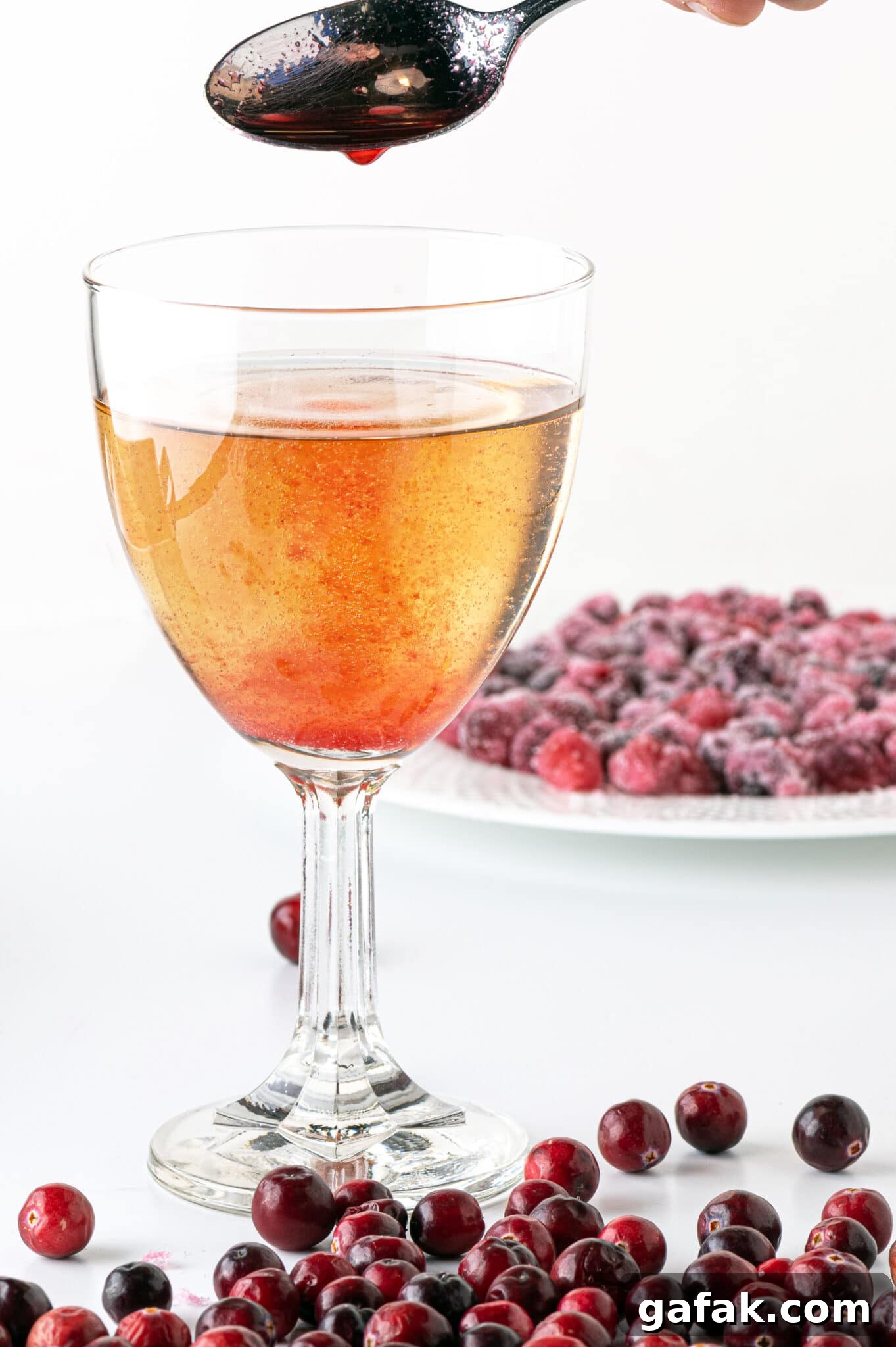 A golden beverage in a wine glass with a stream of cranberry syrup being poured in.   A dish of sugar-free candied cranberries are in the background with fresh ripe cranberries in the foreground. 