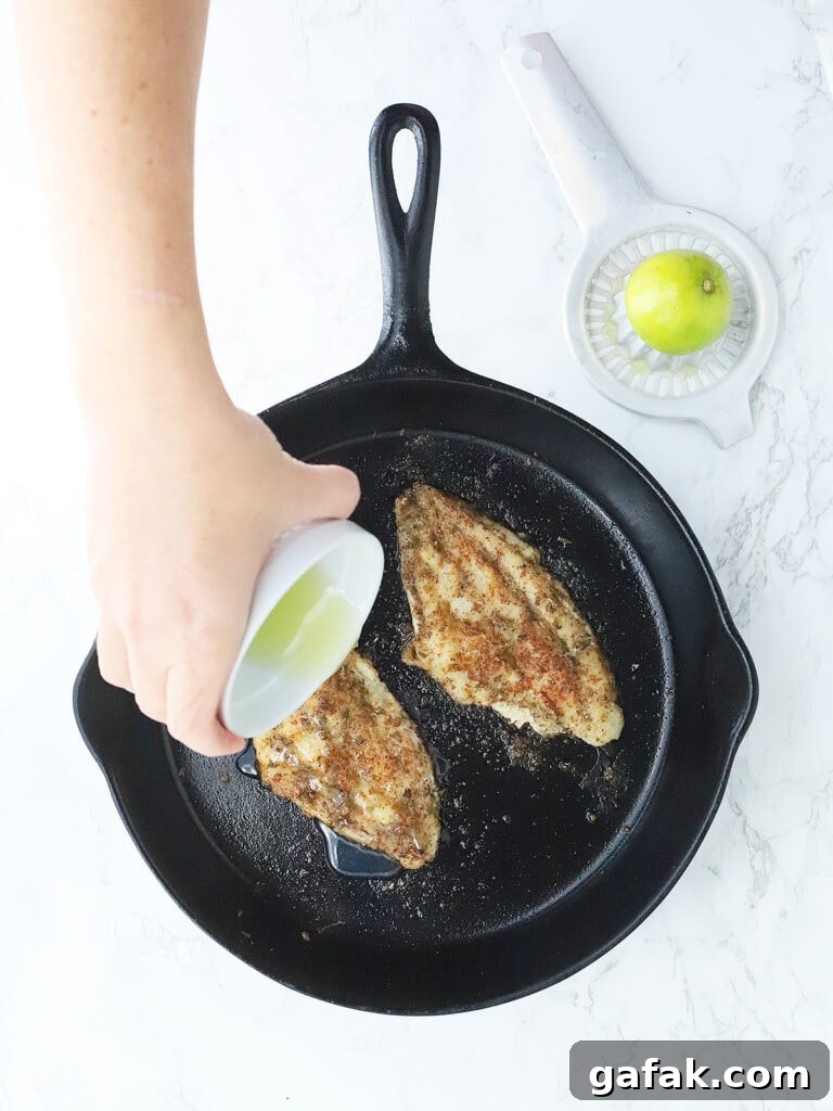 Hand pouring lemon juice over two catfish fillets in a cast iron skillet.