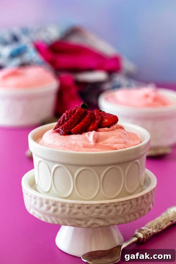 A white ramekin of mousse on a small white pedestal. There's a strawberry fanned on top and two more ramekins of mousse in the background.