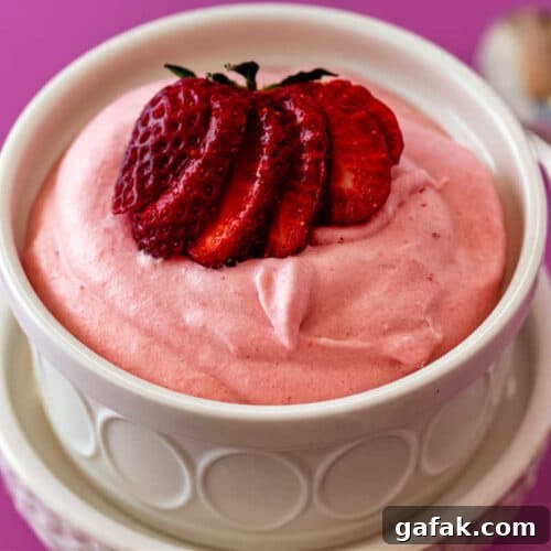 A high-angle shot into a white ramekin of billowy, candy pink strawberry mousse with a cut strawberry fanned on top.