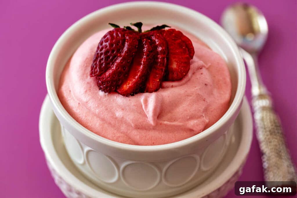A high-angle shot into a white ramekin of billowy, candy pink strawberry mousse with a cut strawberry fanned on top.