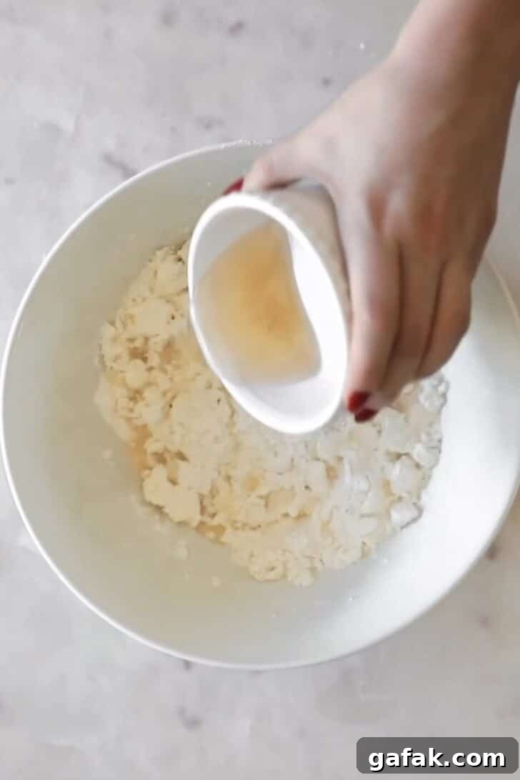 A hand pouring cold water into a white bowl of flour and butter.