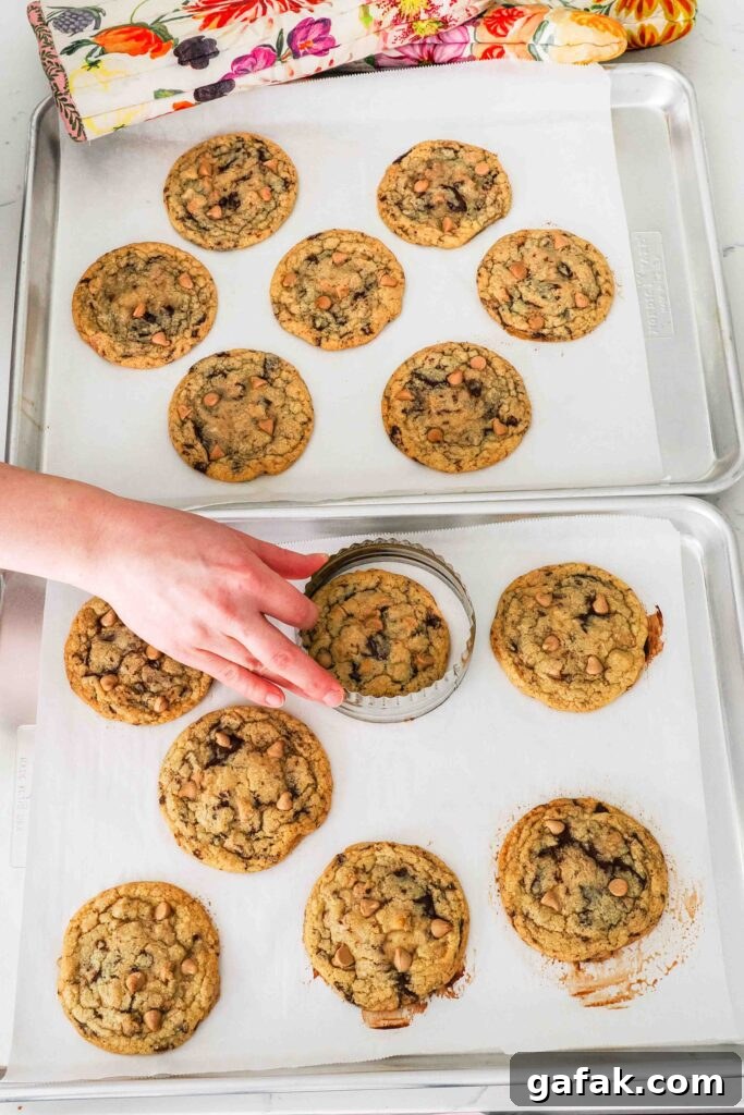 A hand swirls a round cookie cutter around a warm cookie to shape it.