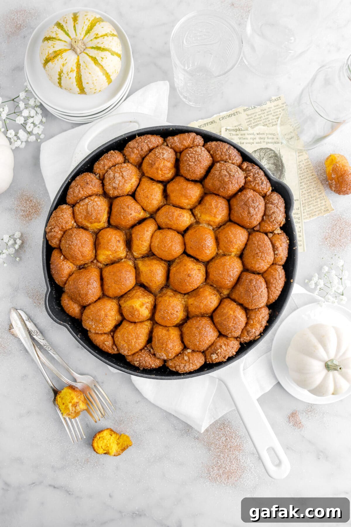 overhead image of skillet pumpkin monkey bread with three forks beside, pumpkins, and white flowers around on marble surface.