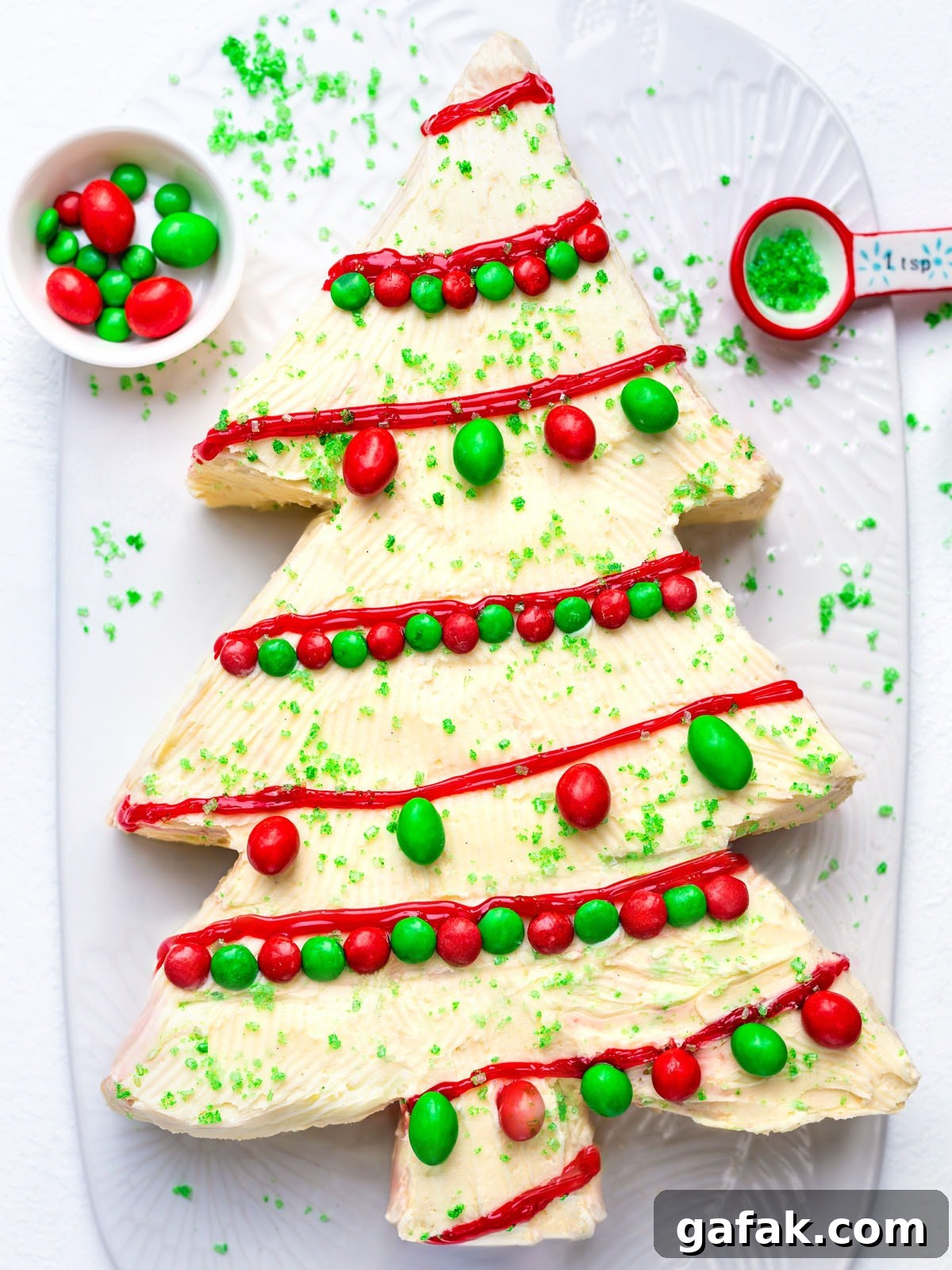 A Christmas tree-shaped cake with white frosting, decorated with red icing, green sugar sprinkles, and red and green candies, sits on a white platter. A small bowl of candies and a teaspoon of sprinkles are nearby.