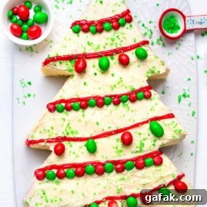 A Christmas tree-shaped cake with white frosting, decorated with red icing, green sugar sprinkles, and red and green candies, sits on a white platter. A small bowl of candies and a teaspoon of sprinkles are nearby.