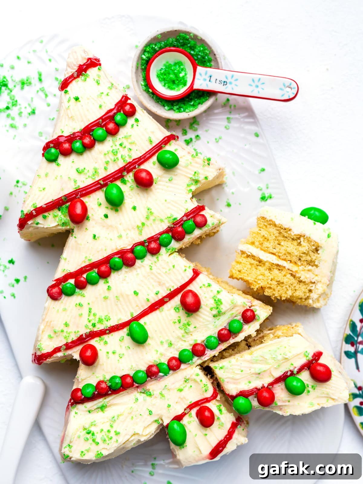 A Christmas tree-shaped cake with white frosting, red icing garlands, green and red candies as ornaments, and green sugar sprinkles. A slice is cut out, and a spoon with green sprinkles is beside the cake.