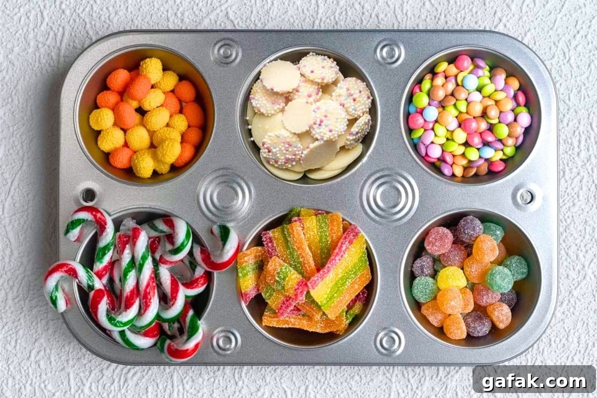 A muffin tray with six compartments filled with various colorful candies, including gummies, candy canes, chocolate buttons, and round sugar-coated sweets, placed on a textured white surface.