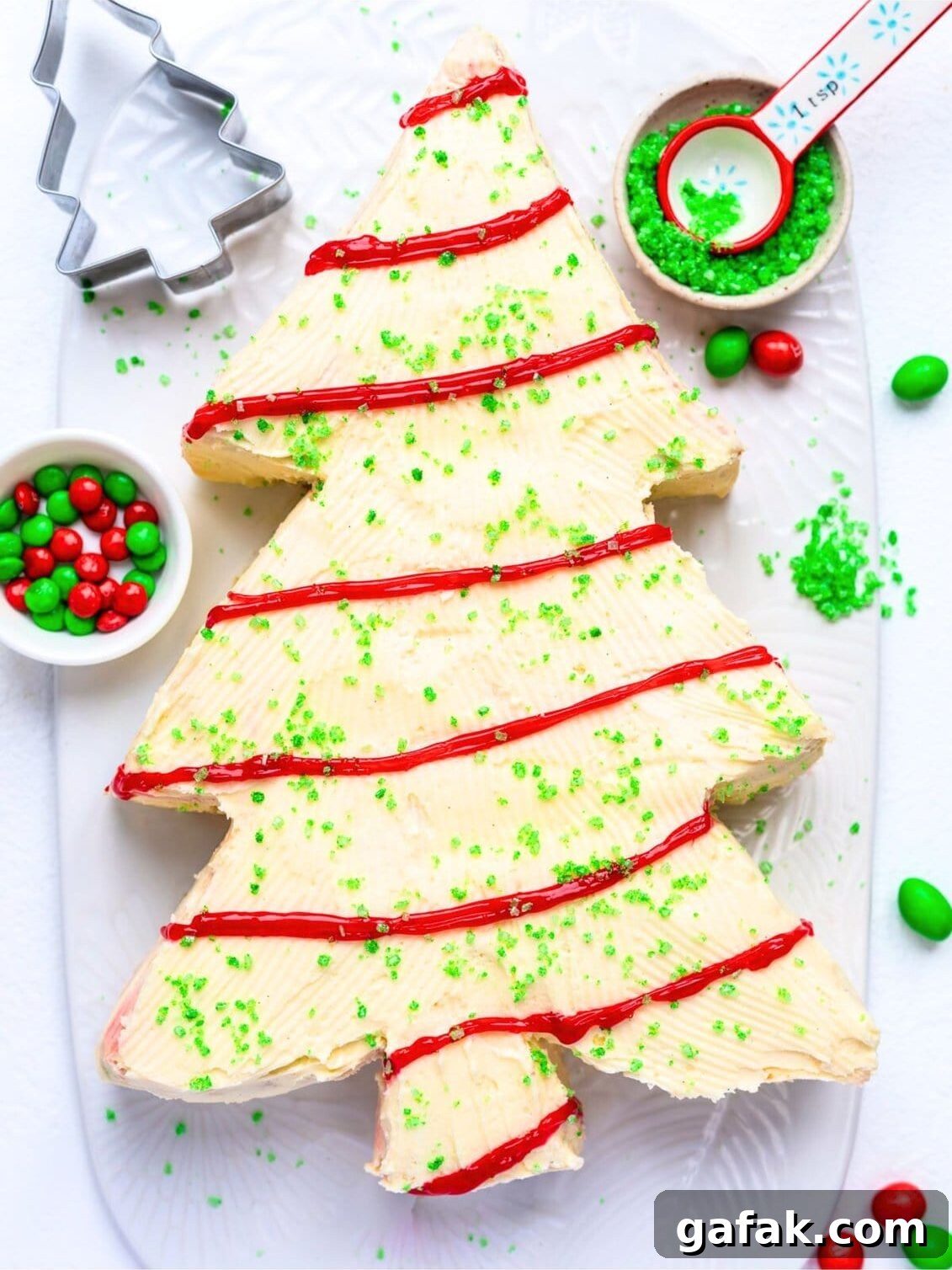 A festive Christmas Tree Cake with white frosting, red icing garlands, and green sprinkles sits on a white platter. Red and green candies, a measuring spoon, and a tree-shaped cookie cutter surround this delightful holiday centerpiece.