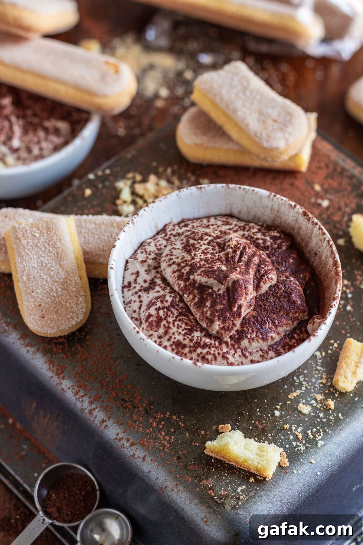 A bowl of tiramisu dessert dip with ladyfingers, cocoa powder and crumbs scattered around.