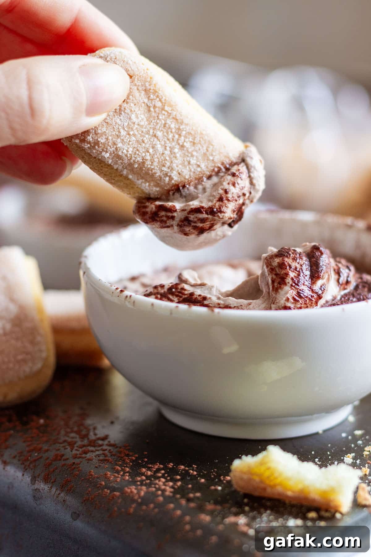 A hand dips a ladyfinger into a bowl of creamy tiramisu sweet dip.
