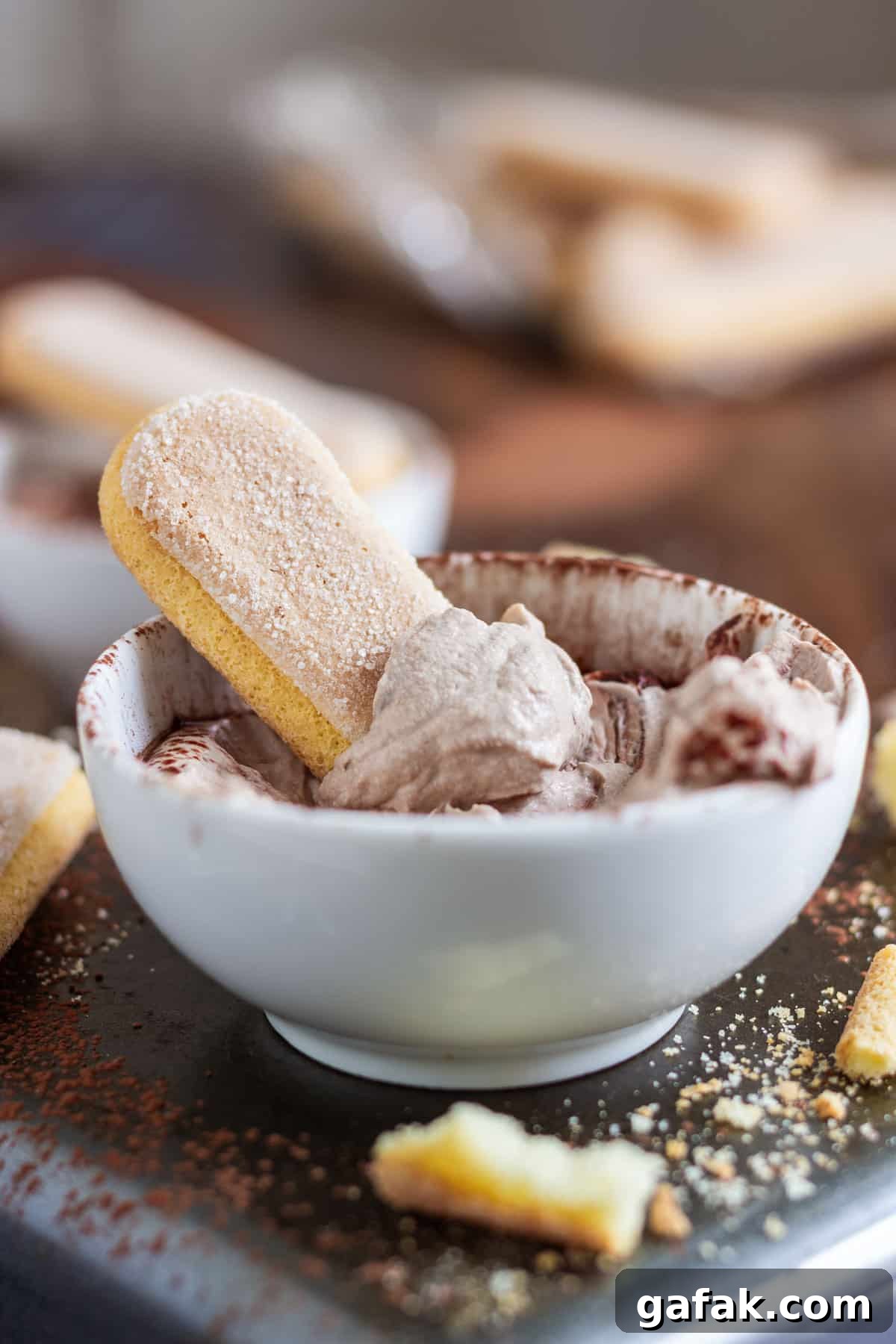 A ladyfinger dipped in coffee-flavored tiramisu dip in a white bowl, with crumbs and more ladyfingers around.