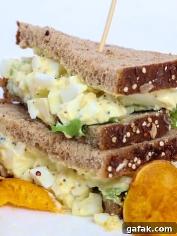 Two halves of an egg salad sandwich stacked, on a plate with sweet potato chips.
