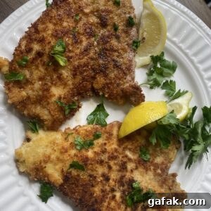 Two pieces of breaded and pan-fried schnitzel on a white plate, garnished with fresh parsley and served with two lemon wedges.
