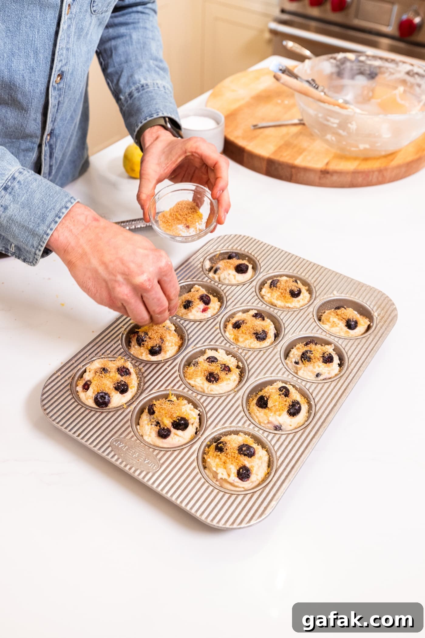 Hand holding small bowl filled with mixture of lemon zest and turbinado sugar sprinkling on top of blueberry muffins before being baked.
