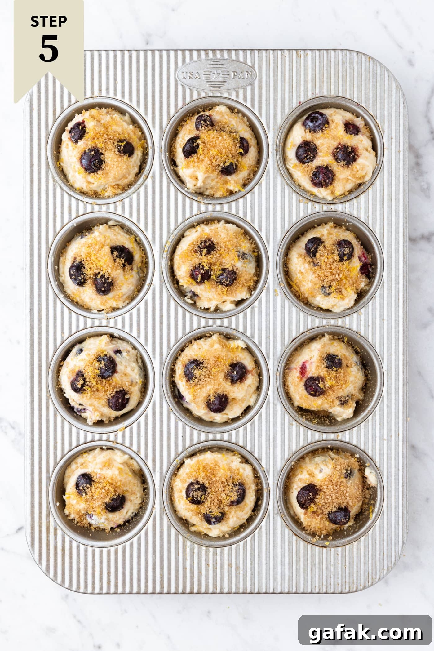 Metal muffin tin sitting on marble surface filled with unbaked batter for blueberry muffins.