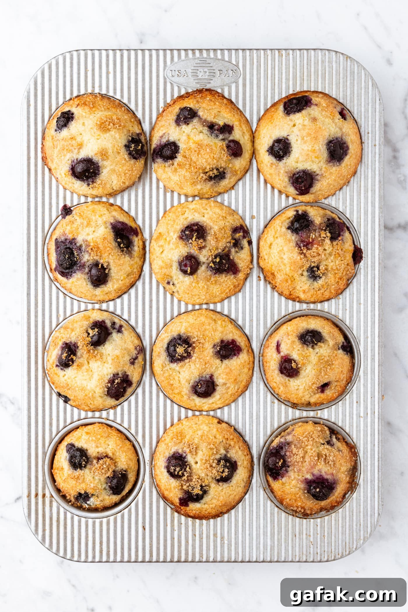 White marble surface with metal muffin tin with freshly baked blueberry muffins with lemon zest topping.
