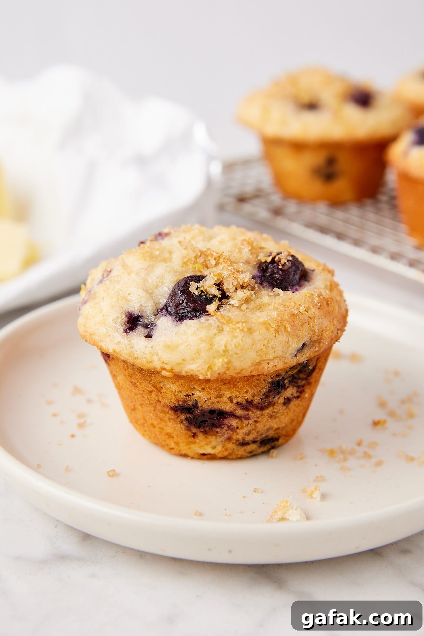 White plate with blueberry muffin with lemon zest turbinado sugar topping with butter in the background.