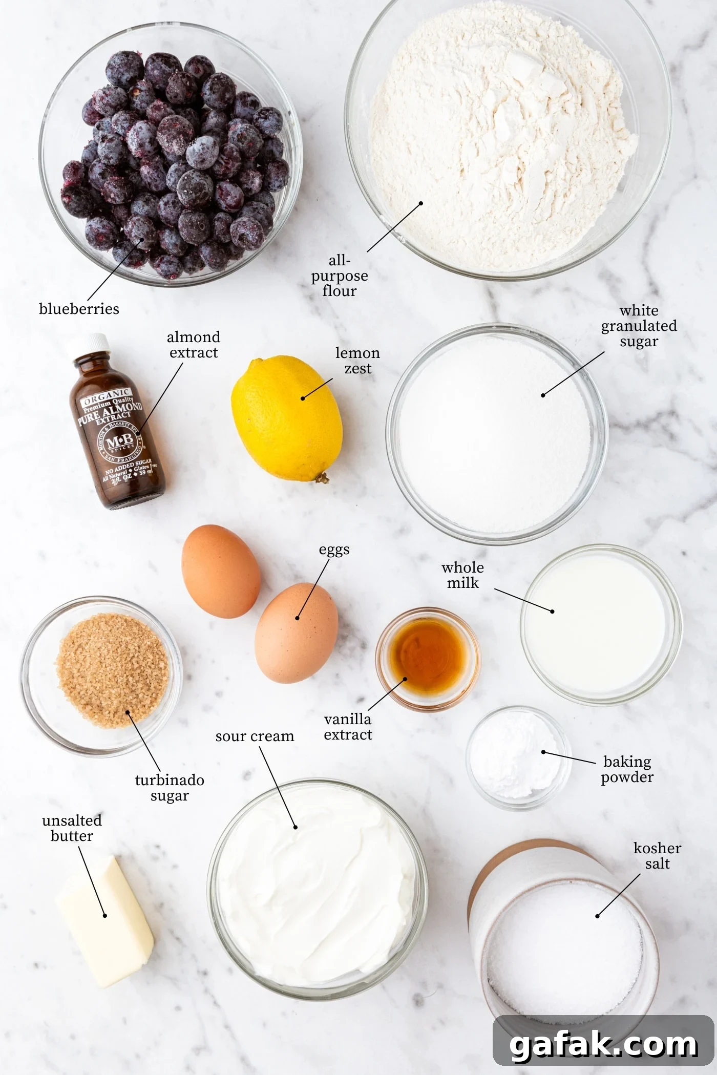 White marble surface filled with all ingredients needed for blueberry muffins including flour, sugar, eggs, spices, blueberries, and more.