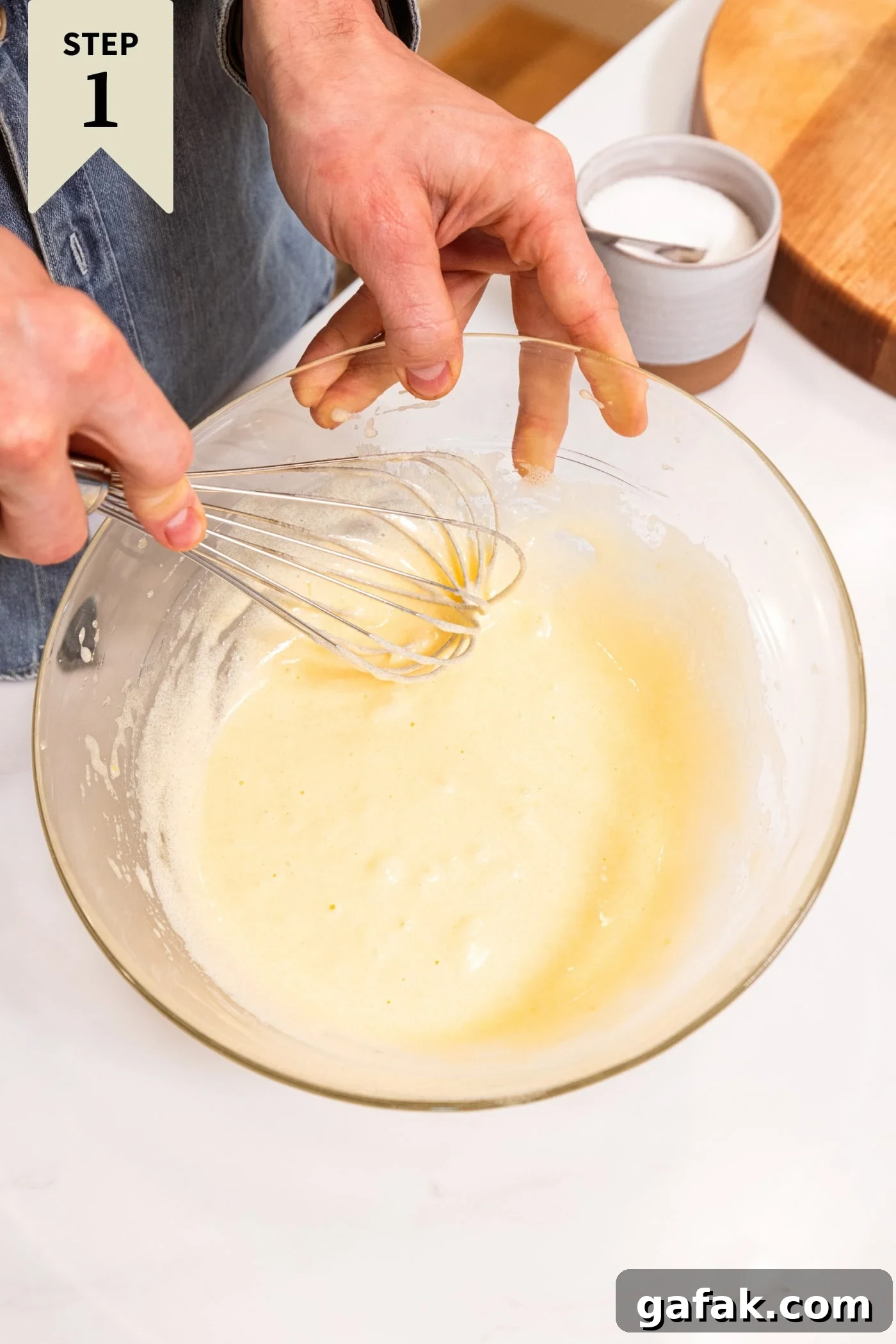 Glass bowl filled with batter for blueberry muffins with whisk whisking everything together all on white surface.