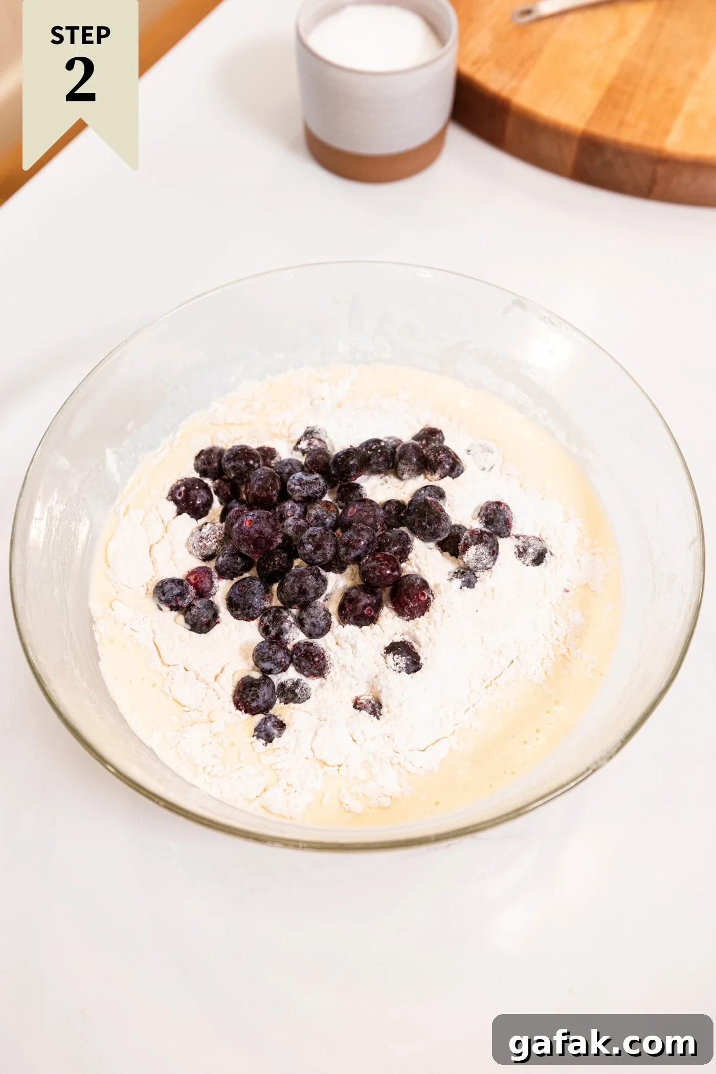 Glass bowl filled with flour on top of batter with frozen blueberries sprinkled on top all for blueberry muffins.