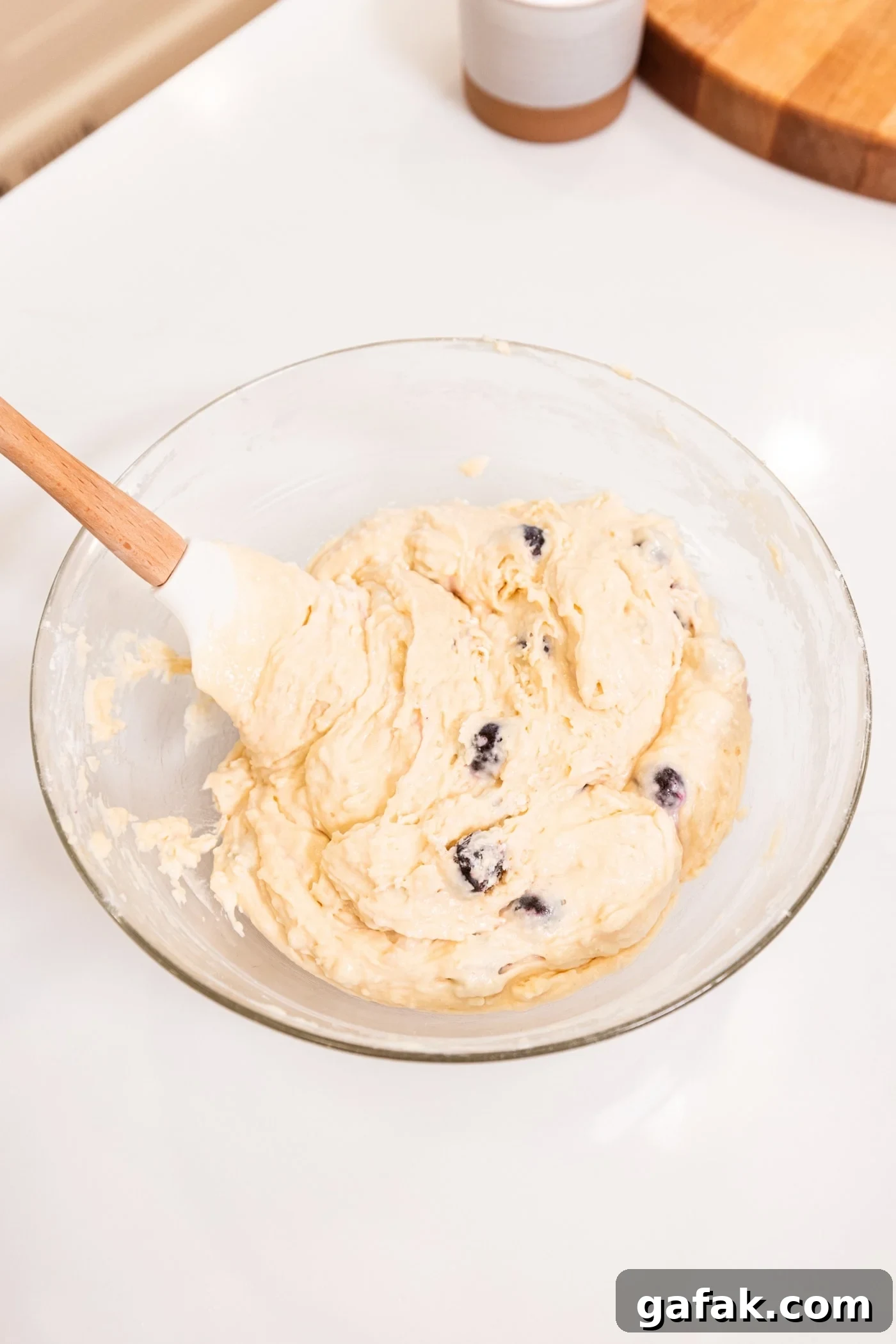Glass bowl filled with batter with blueberries folded in all on white surface.