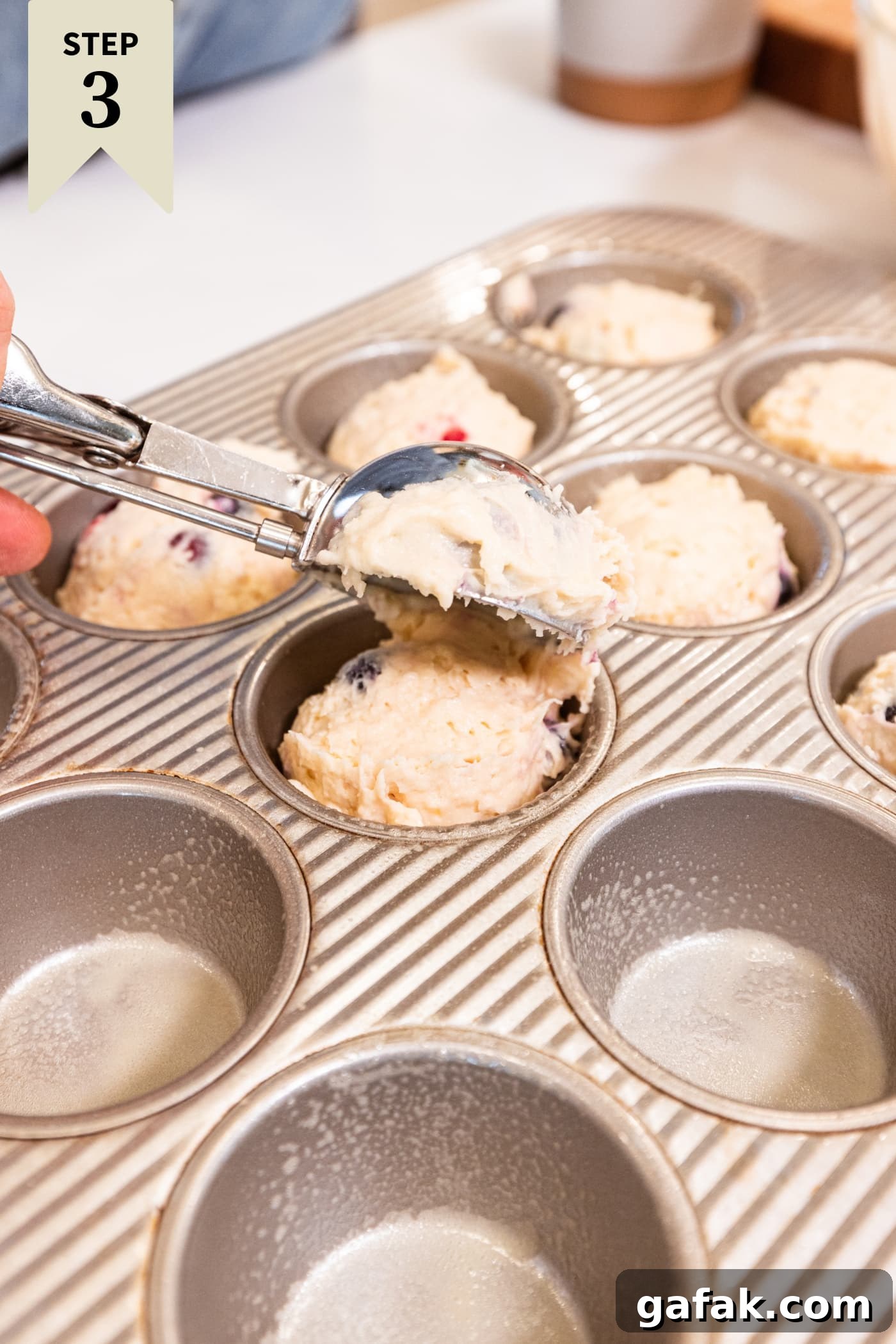 Muffin tins filled with blueberry muffin batter with scoop scooping batter into tins.