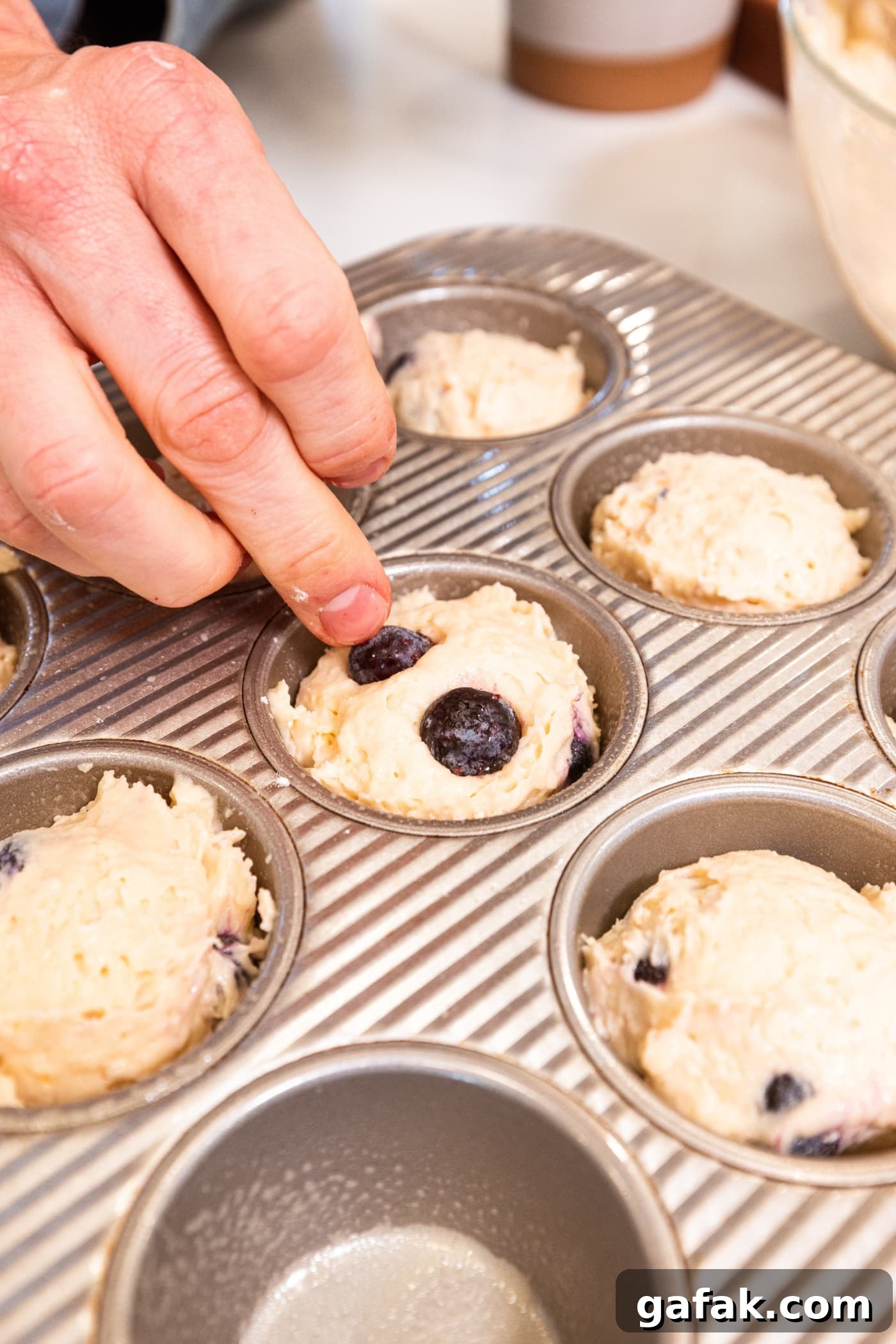 Hand placing blueberries on top of batter in muffin tins for blueberry muffins.