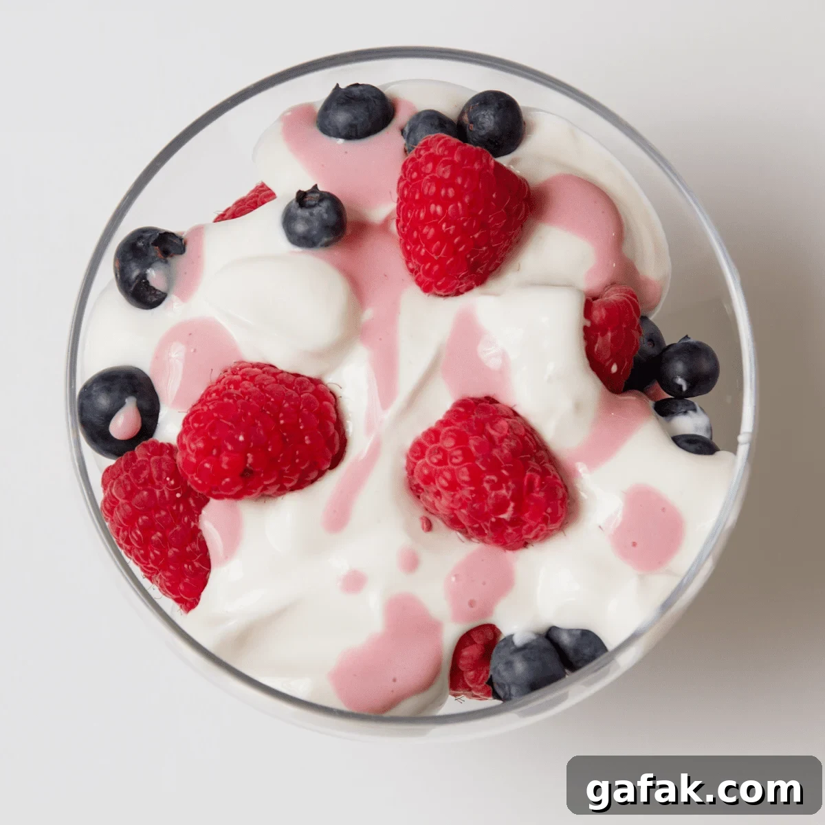 Clear bowl on white table, filled with white yogurt with blueberries, raspberries and pink fruit juice drizzled on top