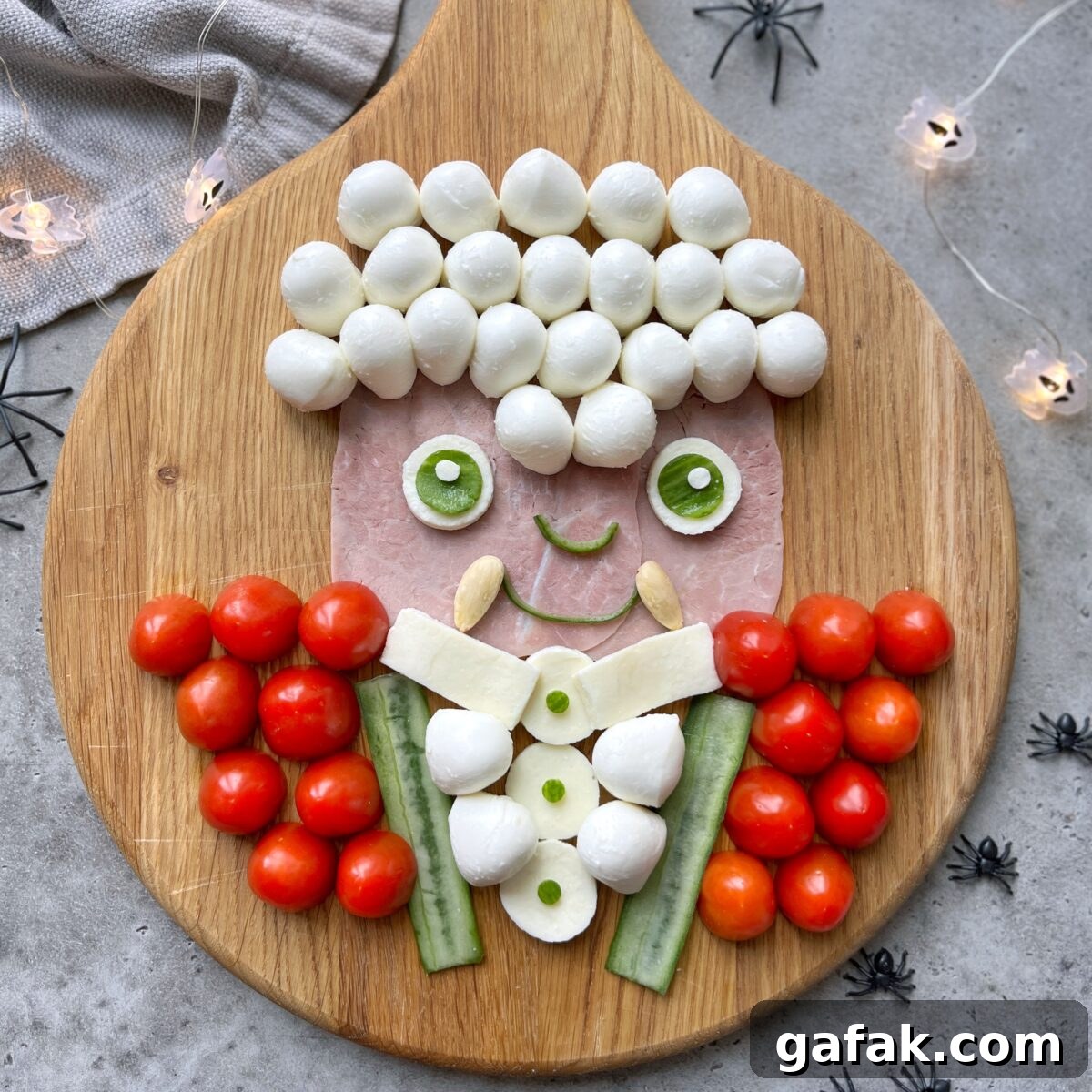 A Vampire face made from ham, mozzarella balls, cucumber, cherry tomatoes, and cheese is arranged on a wooden board. Surrounding it is small plastic spiders and string lights.