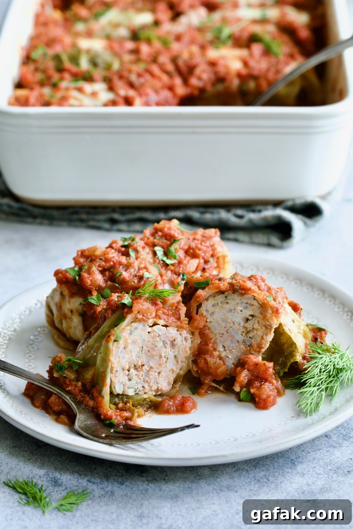 Polish Stuffed Cabbage roll cut in half to expose filling on white plate with baking dish of cabbage in the background.