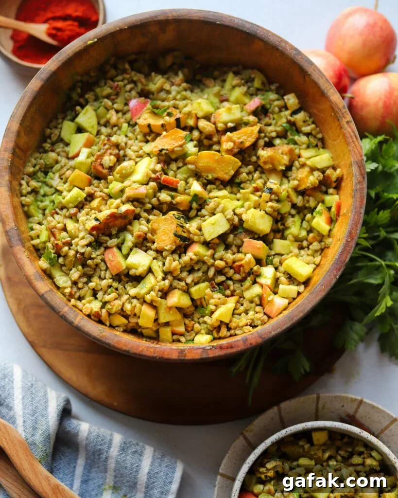 overhead view of a wooden serving bowl with a grain salad made with farro, apple, roasted delicata squash, pecans, and more all tossed in a parsley dijon vinaigrette next to apples and fresh parsley.