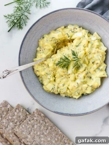 Egg salad in a bowl with a spoon next to crackers and dill sprigs.