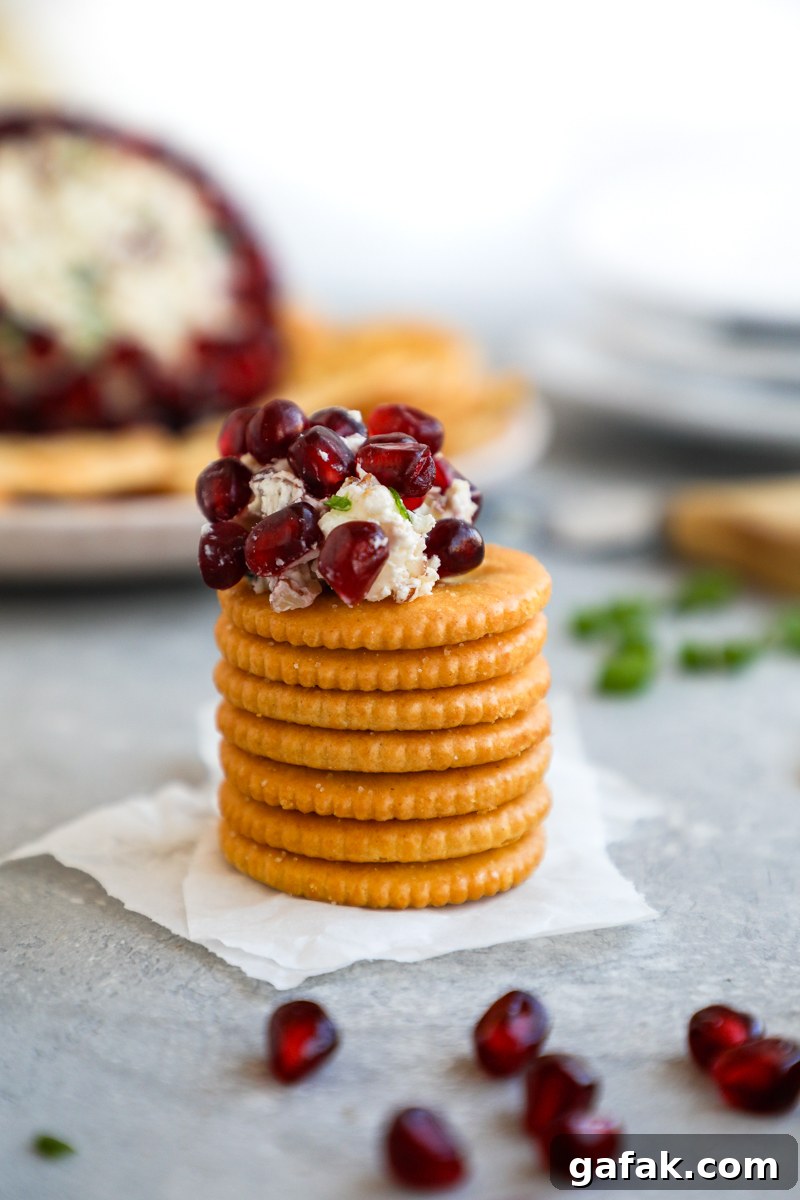 A stack of Ritz crackers with a scoop of a seasonal cheese ball on top with a pomegranate white cheddar cheese ball in the background