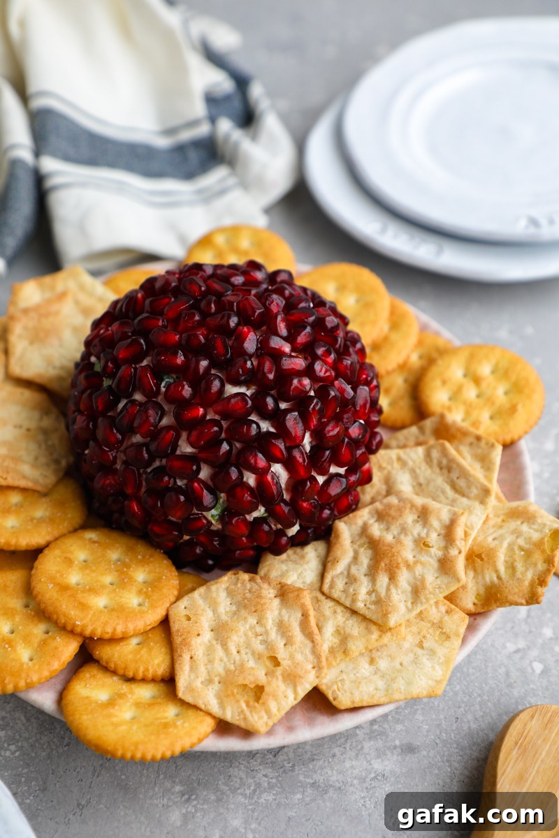 A pomegranate white cheddar cheese ball sitting on a pink plate surrounded by a cheese knife, Rtiz crackers and gluten free crackers