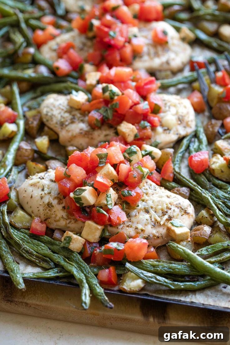 Side closeup of baked chicken breast surrounded by vegetables on pan with Caprese topping cascading across.