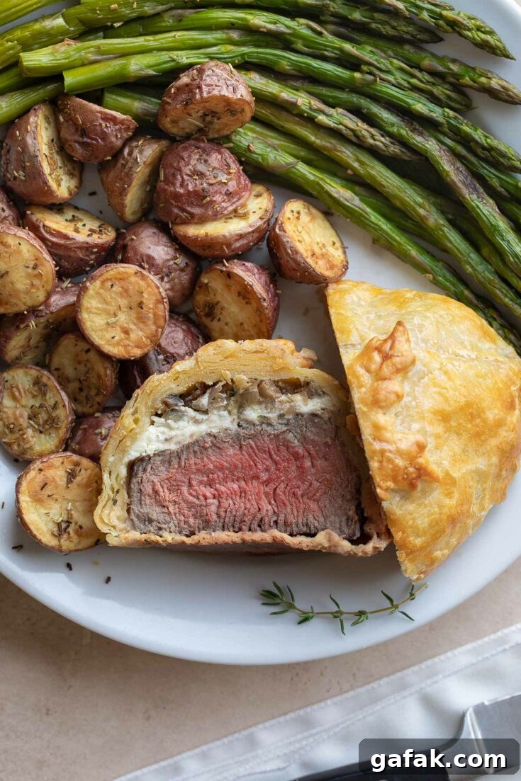 Flatlay of an individual beef Wellington, cut in half with one half facing upwards, surrounded on white plate by roasted asparagus and potatoes.