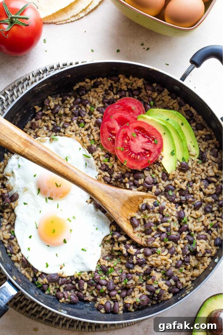 Overhead of cooked Gallo Pinto in pan with minced cilantro, avocado and tomato slices, 2 fried eggs on top.