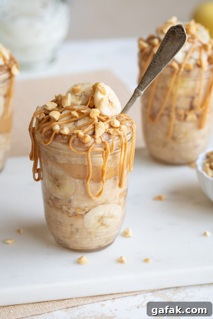 Side closeup of one serving in pretty jar, garnished with sliced bananas and chopped nuts, with peanut butter drizzle and spoon jutting out.