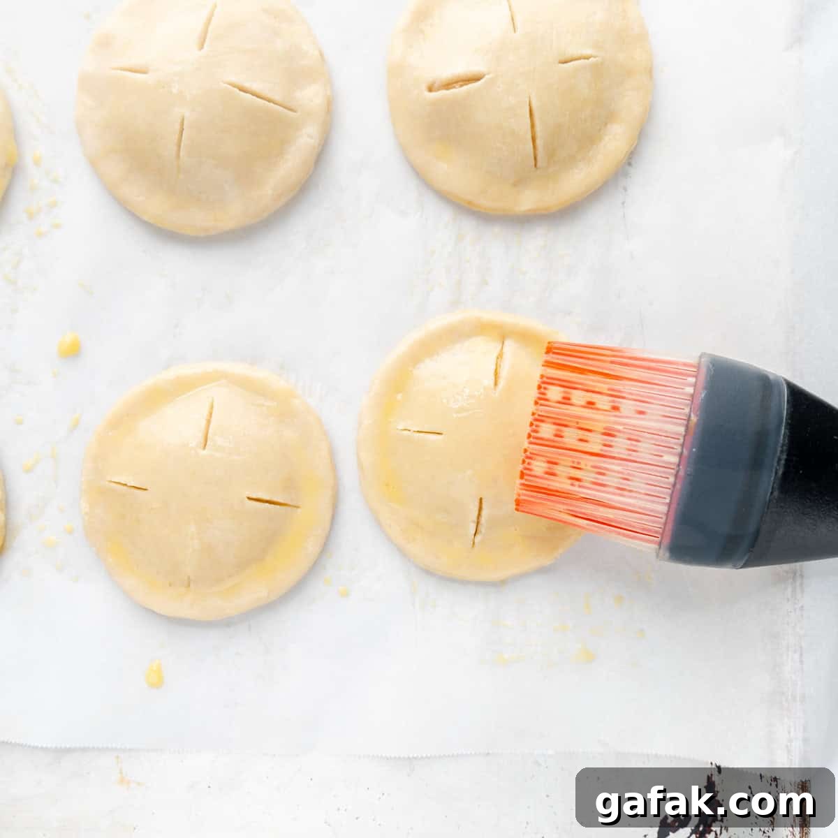 Mini Pumpkin Hand Pies: Flaky Autumn Pastry Recipe 13 A pastry brush glazing pies with egg wash on a white parchment covered pan.