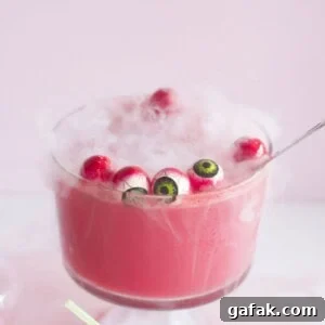 A large glass bowl filled with a pink, frothy drink, topped with floating eyeball decorations and shrouded in mist. Plastic black spiders are scattered around the base. A small glass bottle with a straw sits nearby. Pale pink background.