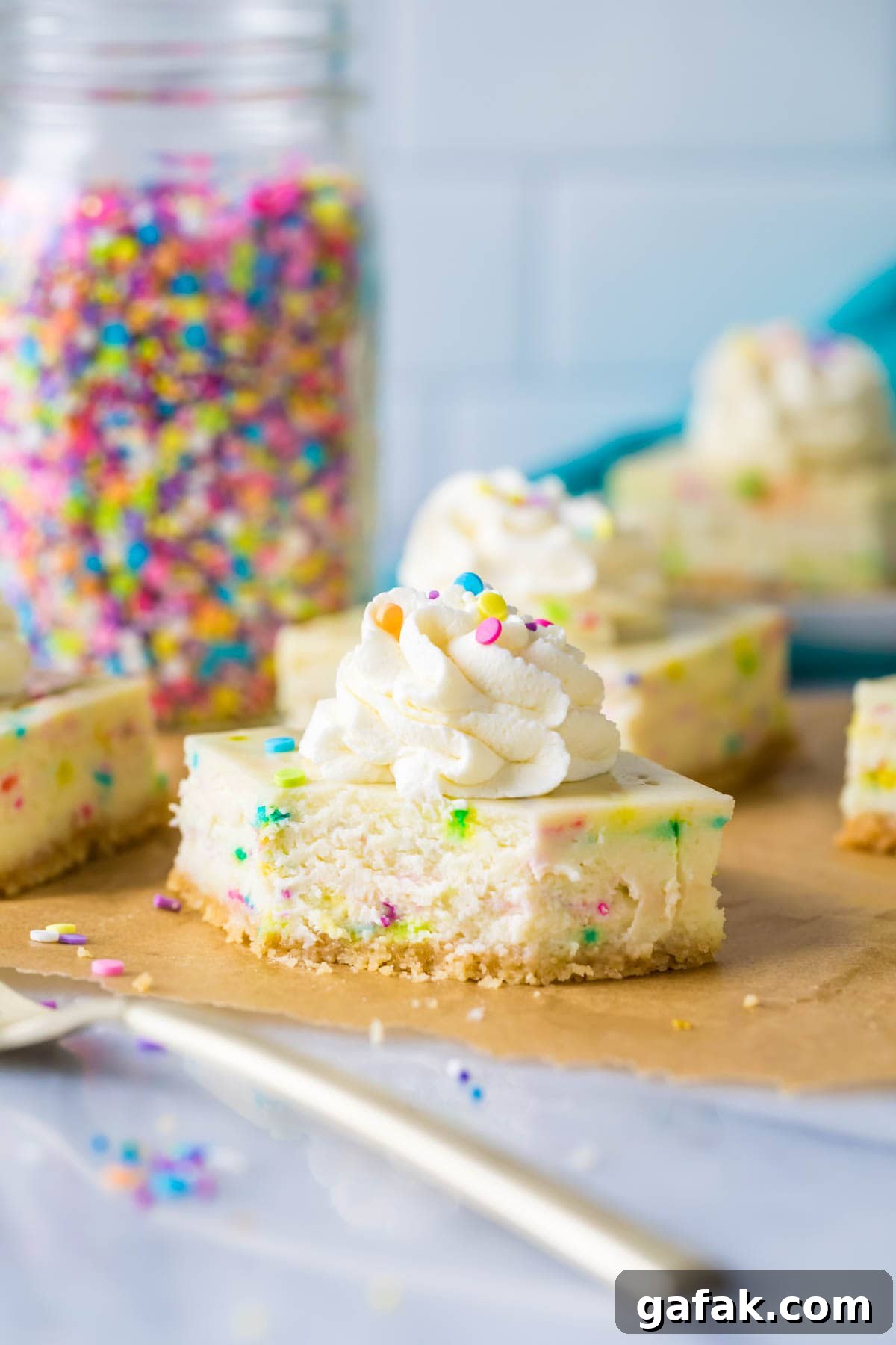 sprinkle-studded cheesecake bar with a bite out of it; gold fork in foreground and jar of sprinkles in background
