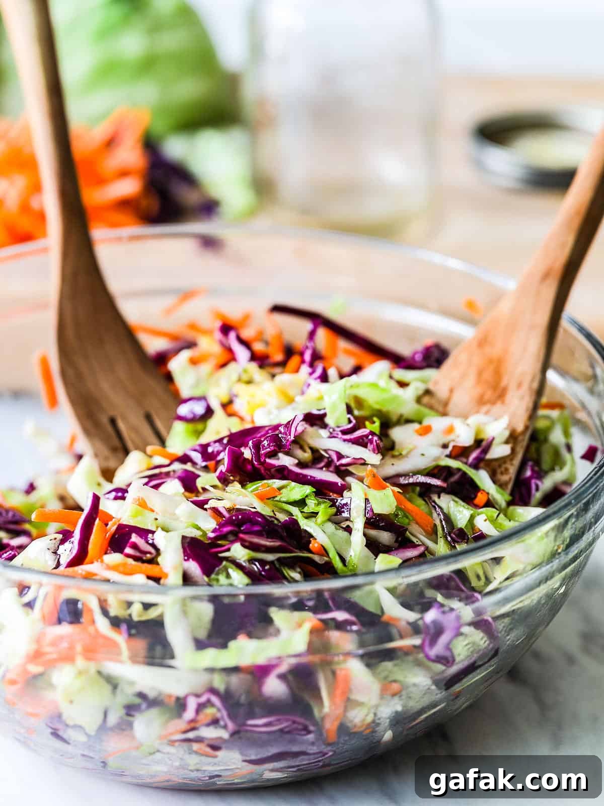 A large clear bowl filled with colorful cole slaw made with purple and green cabbage and carrots with wood serving tongs.
