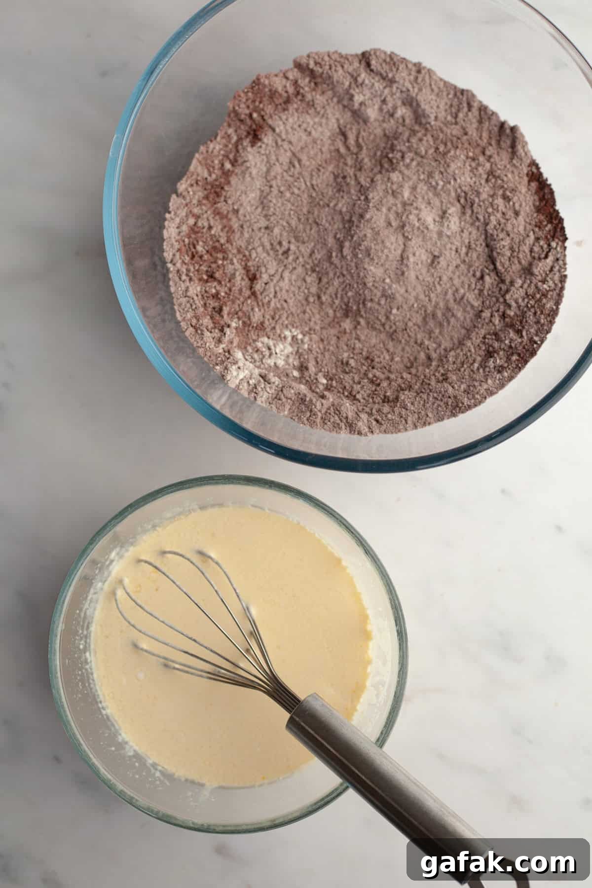 A large bowl with flour and cocoa powder, and a small bowl with a whisk and a pale yellow mixture.